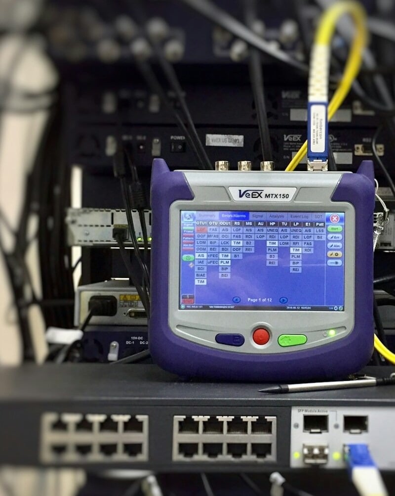 Netzwerk-Switch neben einem Glasfaser-Messgerät auf einem Arbeitstisch.